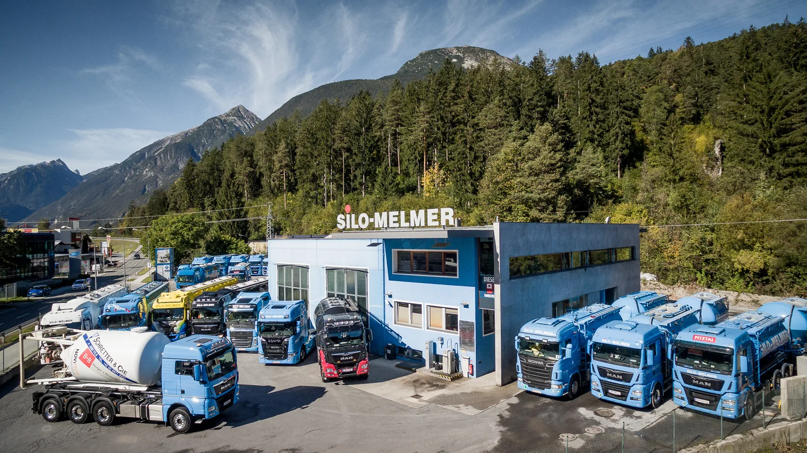 Luftbild der Firmenzentrale von Silo Melmer in Imst: blaues Gebäude mit Schriftzug SILO-MELMER auf dem Dach, davor die LKW-Flotte, dahinter Wald und Berge