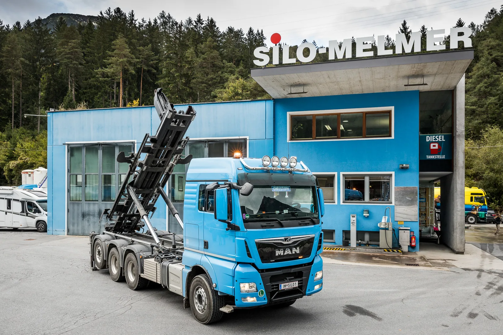 Blauer MAN-Lastwagen mit Abrollkipper vor dem Firmengebäude mit dem Schild Diesel Tankstelle