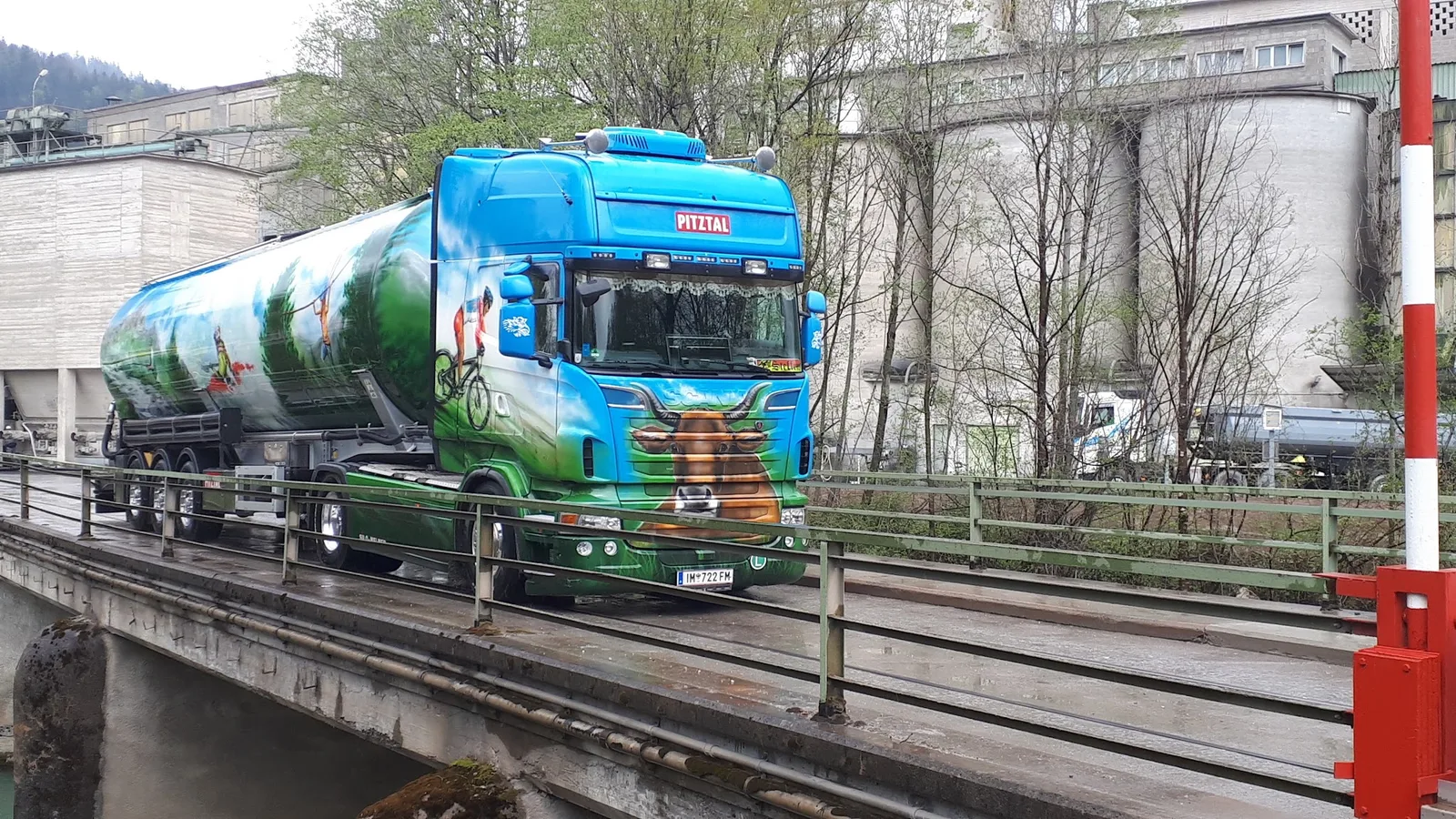 Airbrush-Scania mit Kuhkopf und Radfahrer-Motiv und dem Schild PITZTAL auf einer Brücke vor einem Werk